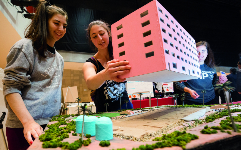 3 students stand around a table that displays a miniature landscape of trees and sand. The middle student holds up a mini paper building.