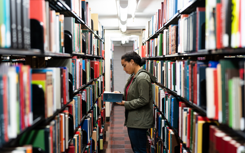 A student reads a book while standing in the stacks. There are two, long bookshelves of books on either side of them.