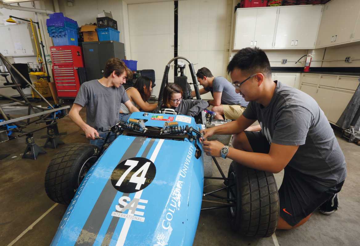 Five students work on a light blue racer car low to the ground.