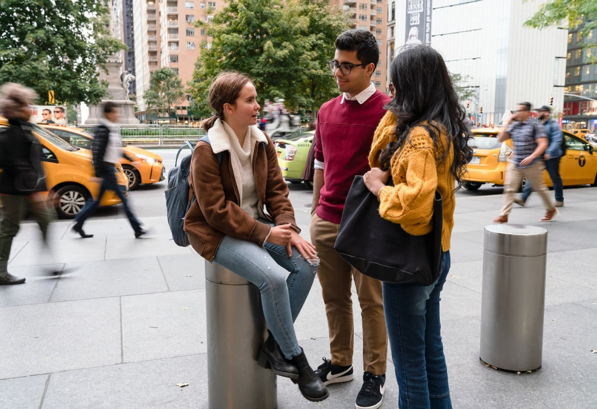Three students sit at a busy intersection as cars and people speed by in the background.