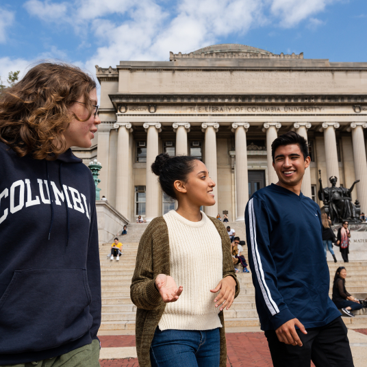 Three students stand in front of Butler library, facing the camera. They are smiling and chatting.