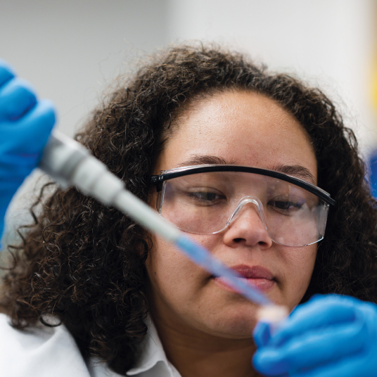 A closeup of a pipette in the foreground with a young female student carefully holding it.