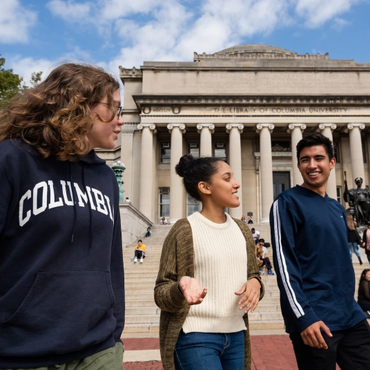 Three students stand in front of Butler library, facing the camera. They are smiling and chatting.