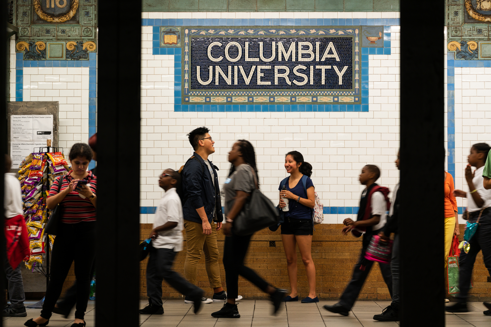 Students and locals walk on a busy subway platform. The tile on the wall reads "Columbia University"