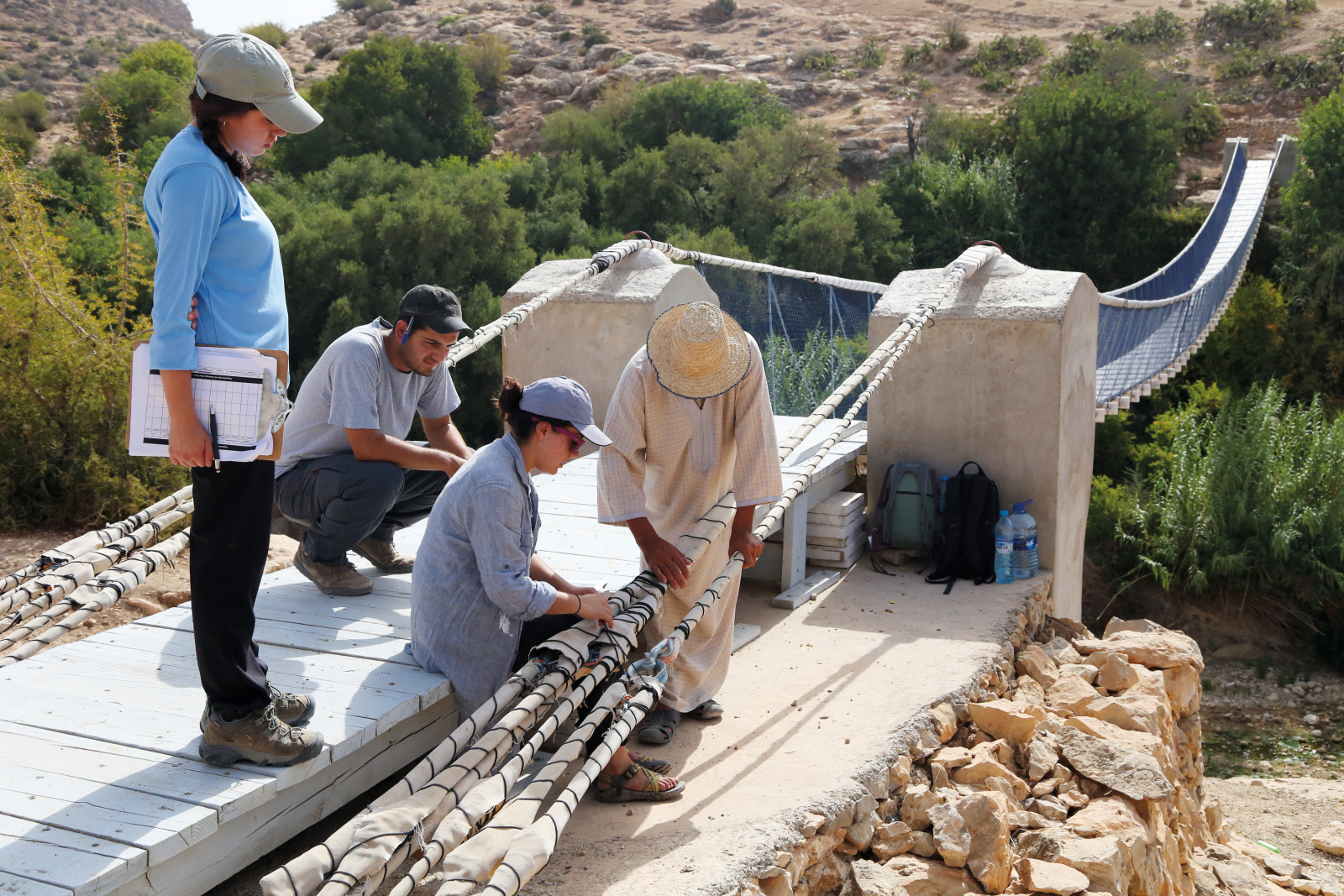 Three students work on a suspension bridge in a desert landscape with a local helper.