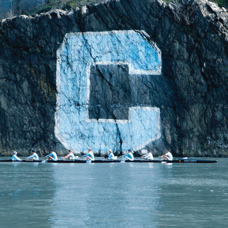 Crew team rowing past rock with large Columbia C