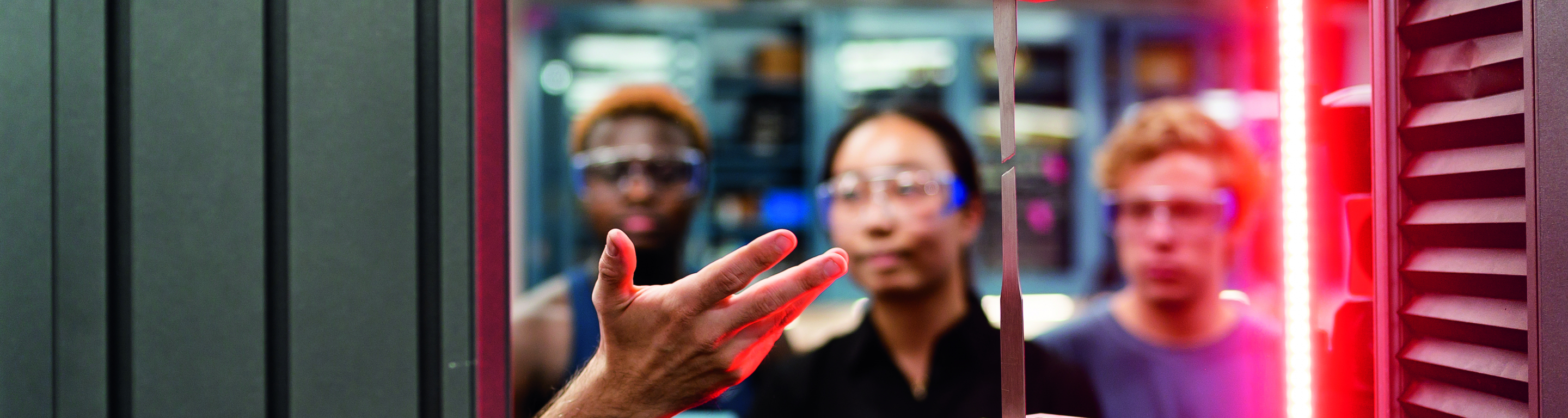 Three students look at a research demonstration, a thin piece of metal is held by two mechanical arms with a laser on the right side of the screen