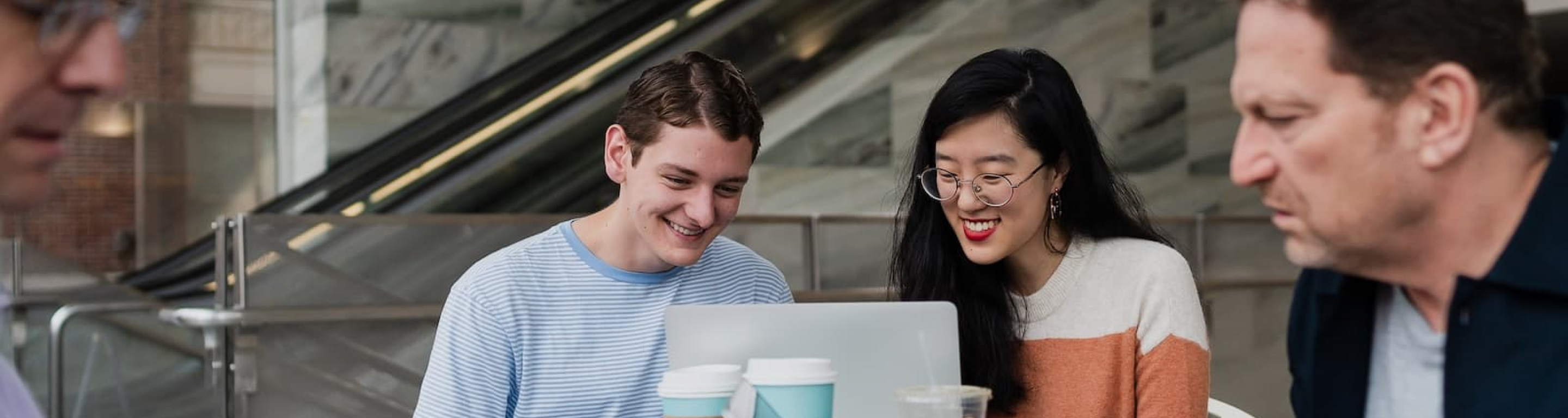 Two students sit at a table, looking at the same laptop and smiling while drinking coffee.