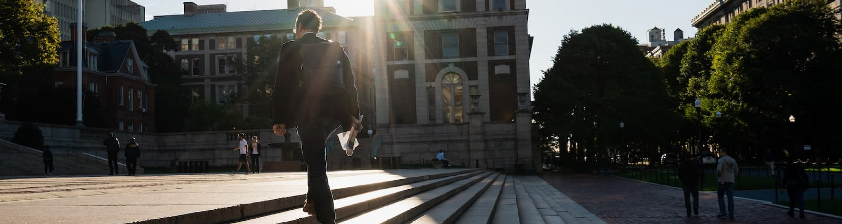 Student dressed in a suit, wearing a backpack and holding a newspaper in their hand, walks up steps at the center of Columbia's campus while bright sunshine streams at him over a rooftop.