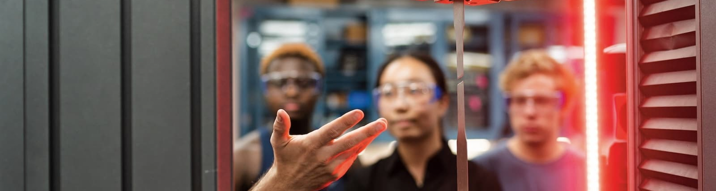 Three students look at a research demonstration, a thin piece of metal is held by two mechanical arms with a laser on the right side of the screen