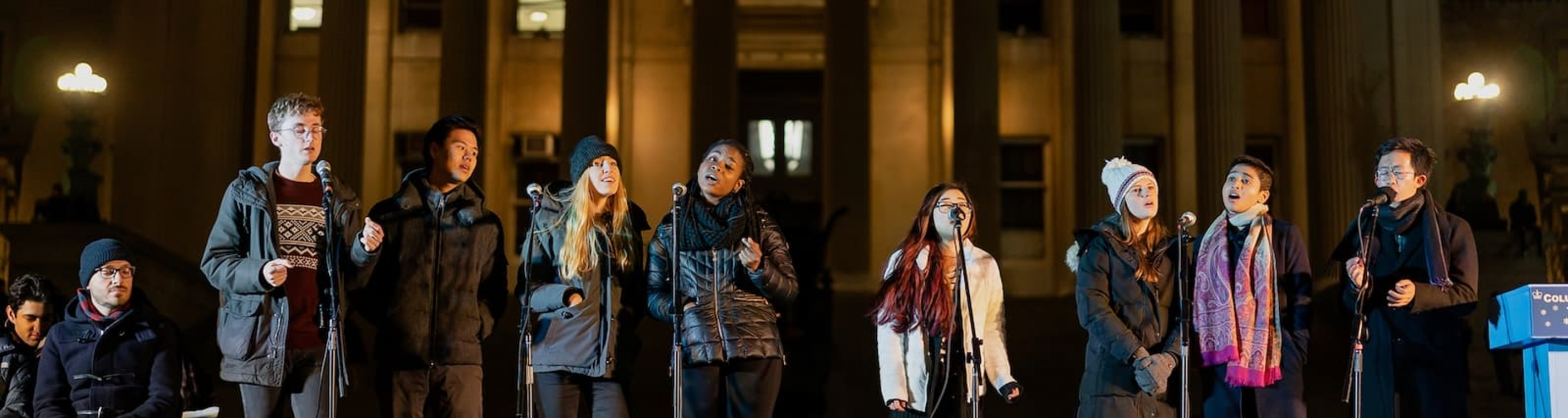 A group of a cappella singers perform on a winter night in front of Low Library. 