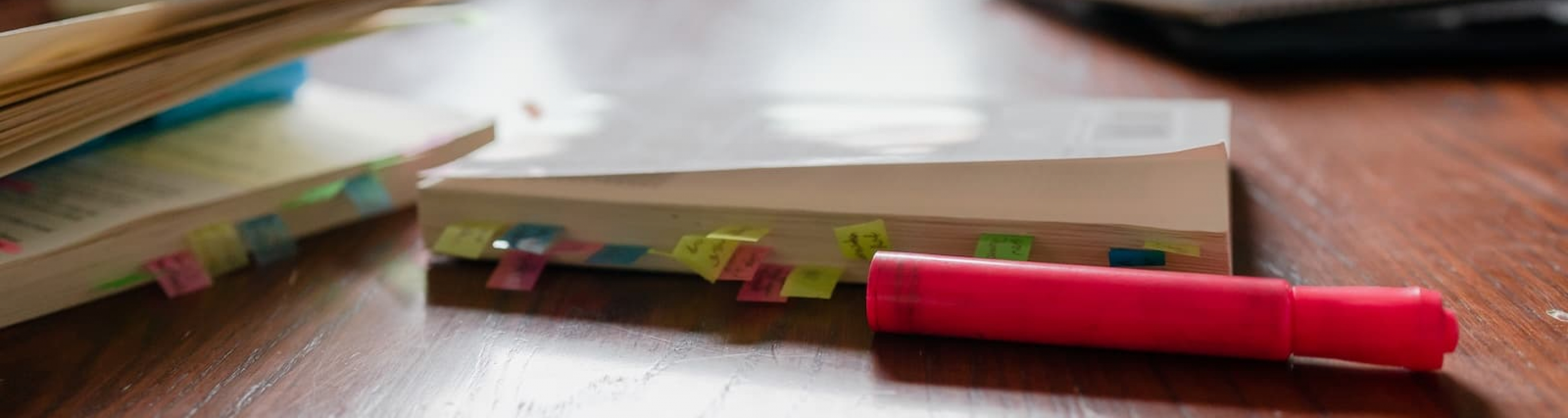 A closeup of a wooden table with a highlighter and multiple books.