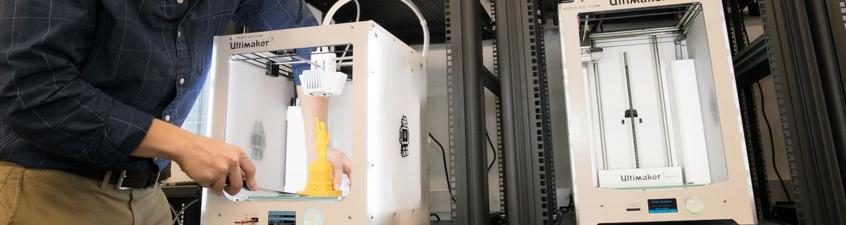 A male student pus away a 3D printer back into a stack of 3D printers in the makerspace.