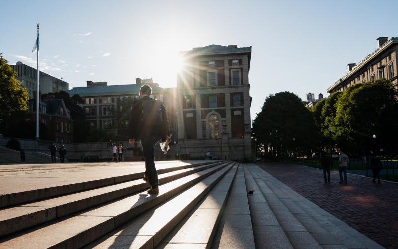 Student dressed in a suit, wearing a backpack and holding a newspaper in their hand, walks up steps at the center of Columbia's campus while bright sunshine streams at him over a rooftop.