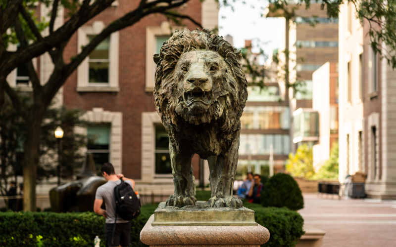 Front view of a bronze lion sculpture with campus buildings in the background.