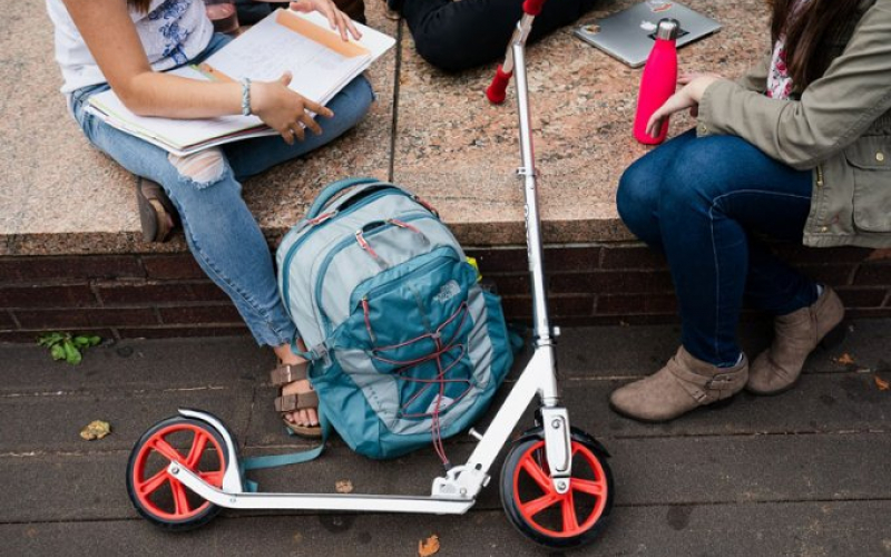 Cropped image of three Columbia students sitting outside with school supplies and a scooter