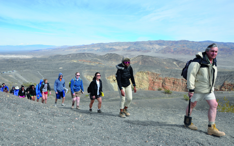 A line of students treks up a rocky, desert path with a professor in front.