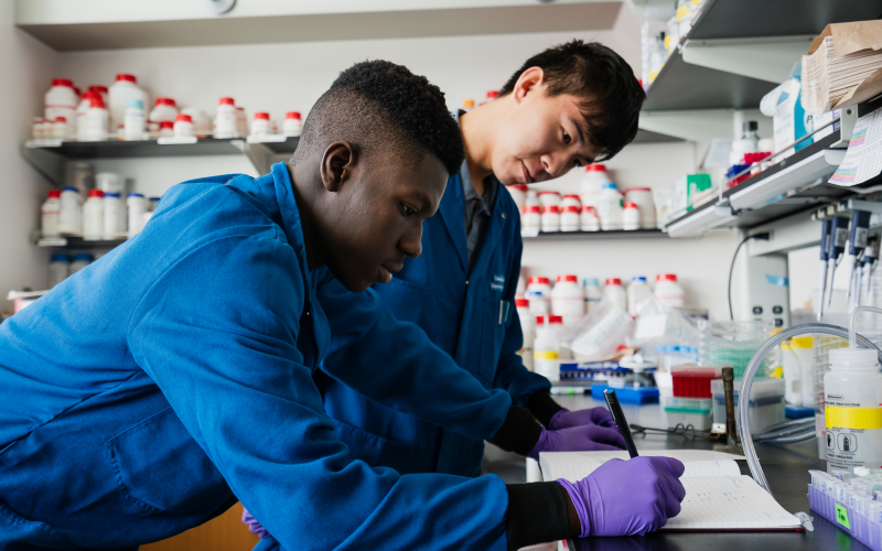 Two students in lab coats and gloves work together in a research lab