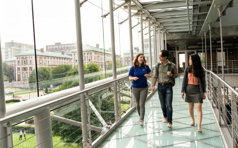 three students walk down a ramp next to a wall of windows showing a view of Columbia's campus