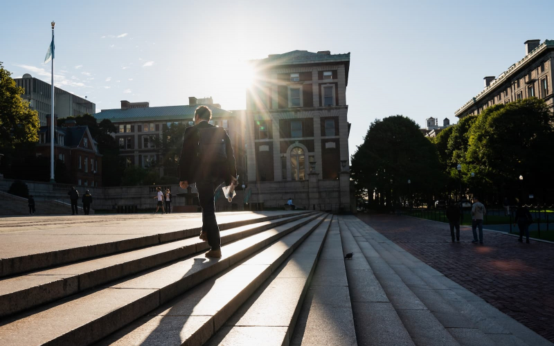 Student dressed in a suit, wearing a backpack and holding a newspaper in their hand, walks up steps at the center of Columbia's campus while bright sunshine streams at him over a rooftop.