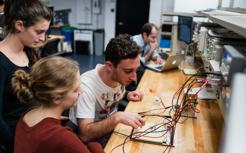 Three students sit at a wooden table and work on an electrical circuit, while smiling.