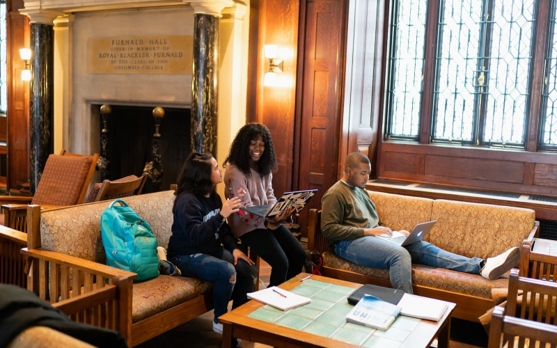 Three students sit in the lounge of Furnald Hall and work on computers and with notebooks