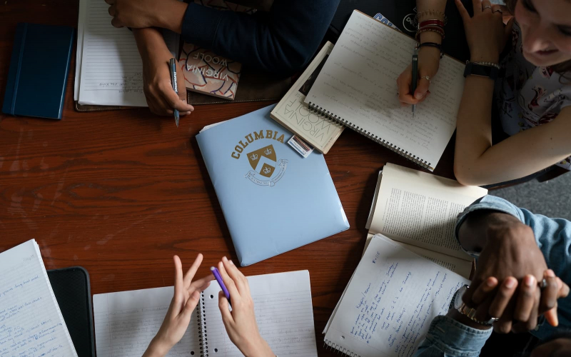 A birds eye view of a classroom table; books, tables, and student hands are scattered.