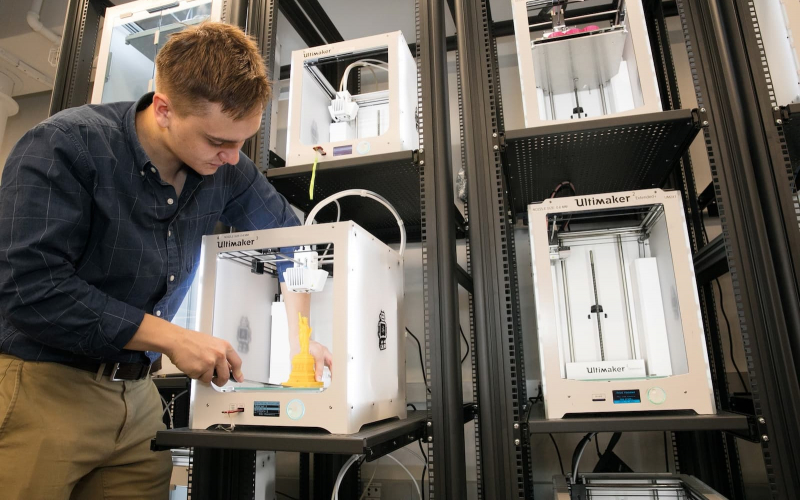 A male student pus away a 3D printer back into a stack of 3D printers in the makerspace.