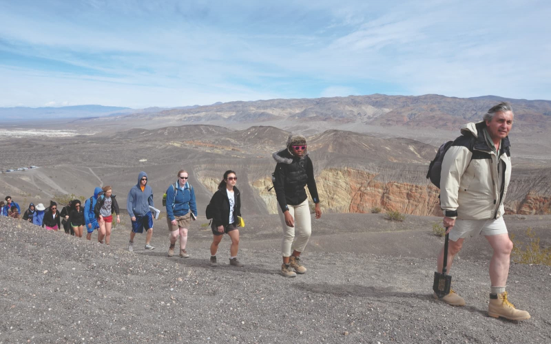 A line of students treks up a rocky, desert path with a professor in front.